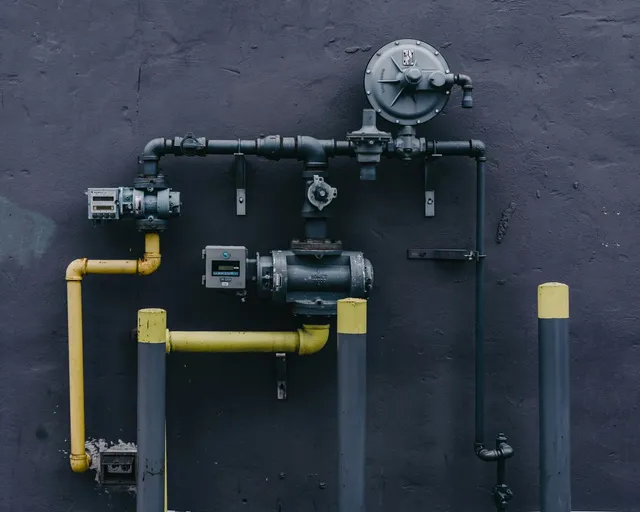 Hand holding HVAC gauges connected to copper pipes; red, blue, and yellow hoses against a brick wall. Red fire hydrant and pipes in front of a building, surrounded by greenery.